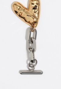 Pendente a forma di cuore in oro con superficie riflettente, attaccato a maglie di catena argentate e una barra orizzontale nella parte inferiore.