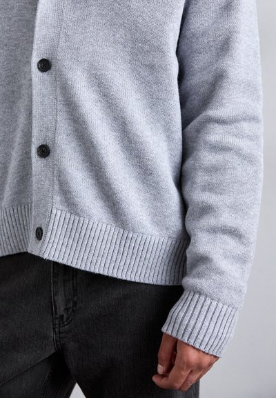 Cardigan lavorato a maglia grigio chiaro con orlo e polsini a costine, caratterizzato da cinque bottoni neri sul davanti. Ingrandimento della texture del tessuto visibile.