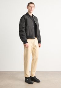 Veste bomber noire avec des poignets et un col côtelés, chinos beiges et baskets noires. Tissu lisse avec des boutons-pression et des poches latérales.