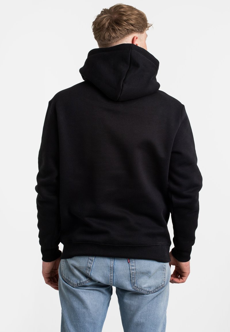 Schwarzer Kapuzenpullover mit Kängurutasche, weichem Stoff, gerippten Bündchen und Saum; getragen mit hellblauen Jeans, die eine lässige Rückansicht zeigen.