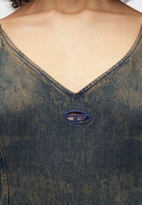 Peça de vestuário em denim com um decote em V profundo, apresentando um recorte oval azul como detalhe. O tecido exibe um efeito desbotado com uma aparência texturizada e gasta.