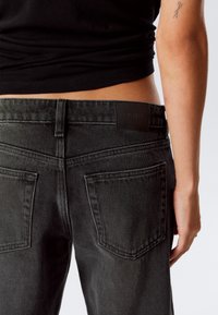Primo piano della parte bassa della schiena di una persona che indossa jeans di denim neri e un top nero cropped, con un piccolo tatuaggio sull'avambraccio e la mano laterale visibile.