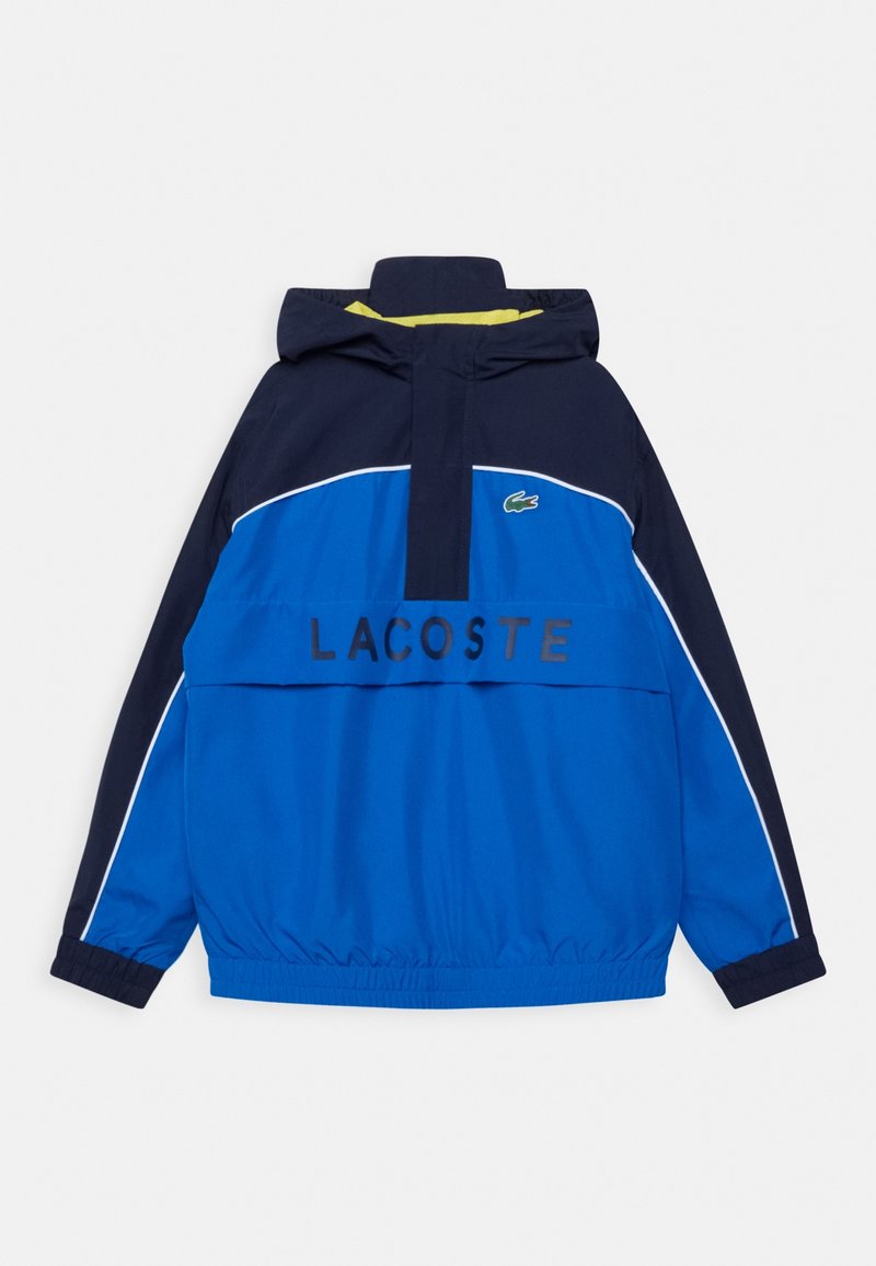 Blue and navy hooded Lacoste jacket with white piping, elastic cuffs, and large front pocket with "LACOSTE" text and crocodile logo.