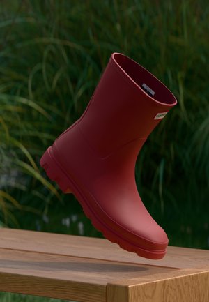 Roter Gummistiefel für den Knöchel mit profiliertem Absatz, gebrandetes Etikett nahe der Oberseite, schwebt über einer Holzbank im Freien mit grünem Gras im Hintergrund.