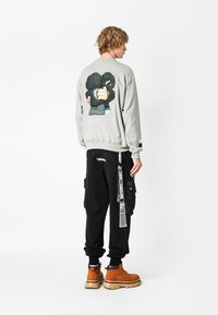 Sudadera gris con un gráfico de dibujos animados en la espalda, combinada con pantalones cargo negros que tienen un bolsillo lateral y botas marrones.