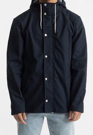 Homme portant une veste à capuche bleu marine avec des cordons blancs et des boutons-pression argentés, associée à un jean bleu clair.