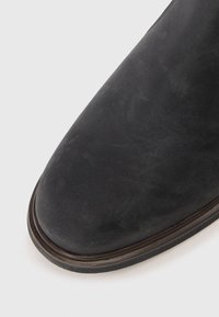 Botine negre din suede cu vârful rotund, textură netedă și o margine din piele maro de-a lungul tălpii.