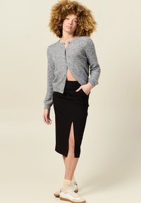 Grau gestrickte Strickjacke mit Knopfverschluss, kombiniert mit einem schwarzen Rock in mittlerer Länge mit seitlichem Schlitz und weißen Sneakers mit beigen Akzenten.