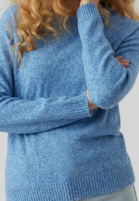 Kvinna med lockigt hår iförd en blå stickad tröja, med armarna i kors och en hand vilande nära hakan mot en enfärgad bakgrund.