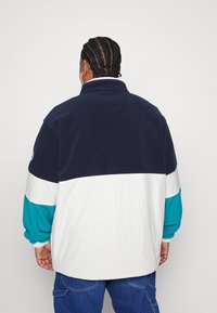 Kurtka polarowa z granatową górą, białym środkowym pasem i turkusowymi dolnymi rękawami. Wysoki kołnierz i luźny krój z elastycznymi mankietami.