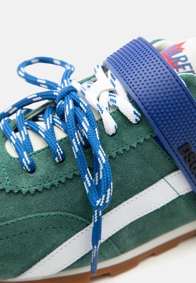 Baskets vertes et blanches avec des lacets bleu et blanc attachés en nœud, avec une sangle bleue texturée sur le dessus et une semelle marron.