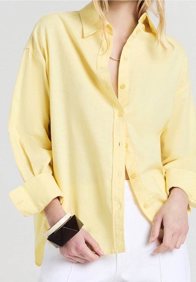 Personne portant une chemise jaune ample boutonnée et un pantalon blanc avec des bracelets géométriques noirs et blancs au poignet gauche.