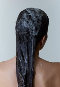 Langt, mørkt hår dekket av hvit, skummende sjampo, sett bakfra, med synlig hud på skuldre og nakke. Myk tekstur og blank finish.