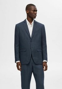 Abito tailored blu navy con giacca monopetto e pantaloni a gamba dritta. Presenta un tessuto testurizzato e revers a punta. Camicia bianca sotto.