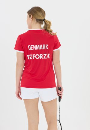 Kvinde med hestehale iført rød "DENMARK FORZA" sportstrøje og hvide shorts, der holder en badmintonketcher og står mod en ensfarvet baggrund.