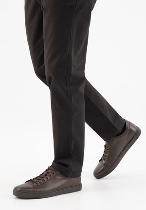 Benedenbenen met zwarte broek en donkerbruine leren sneakers met veters, één voet lichtjes opgetild alsof ze aan het lopen of vooruitstappen zijn.