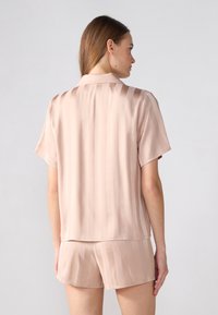 Siden pyjamasset i ljusrosa, bestående av en kortärmad skjorta med ränder och matchande shorts, slät textur och avslappnad passform.