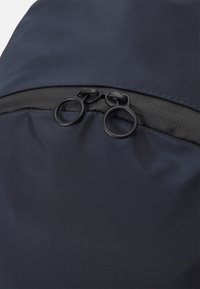 Un sac à dos bleu marine en tissu avec une texture lisse et deux tirettes de zipper circulaires en noir. Les contours du zipper sont discrets par rapport au matériau.