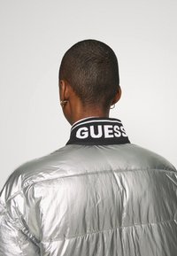 Sølv pufferjakke med en ribbet sort krave, der har "GUESS" i hvid. Tekstureret overflade med en refleksiv finish, og sømmene er synlige.