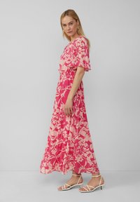 Robe maxi rose avec un motif floral, manches courtes volantées, taille smockée, design fluide, associée à des sandales blanches à lanières.
