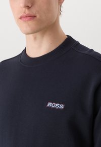 Sudadera azul marino hecha de tejido suave, con un cuello redondo acanalado y el logotipo "BOSS" bordado en azul claro y rojo.