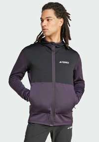 Giacca Terrex nera e viola con zip frontale, cappuccio, pannello superiore texturizzato, pannello inferiore liscio e logo sul petto.
