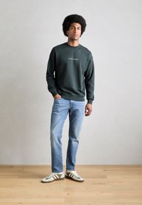 Donkergroen sweatshirt met een wit logo, lichtblauwe spijkerbroek en groen/crème sneakers. Model staat op een houten vloer tegen een grijze muur.
