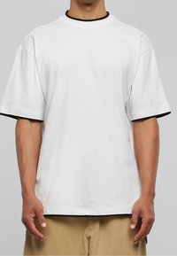 Muž oblečený v bílém oversized tričku přes černou dlouhou košili s khaki kalhotami, stojící na šedém pozadí.