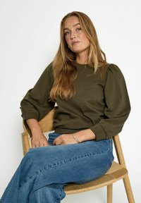 Oliwkowa, długi rękaw, bluzka z bufiastymi ramionami, w połączeniu z jasnoniebieskimi dżinsami. Model siedzi na drewnianym krześle, ręce spoczywają na kolanach.