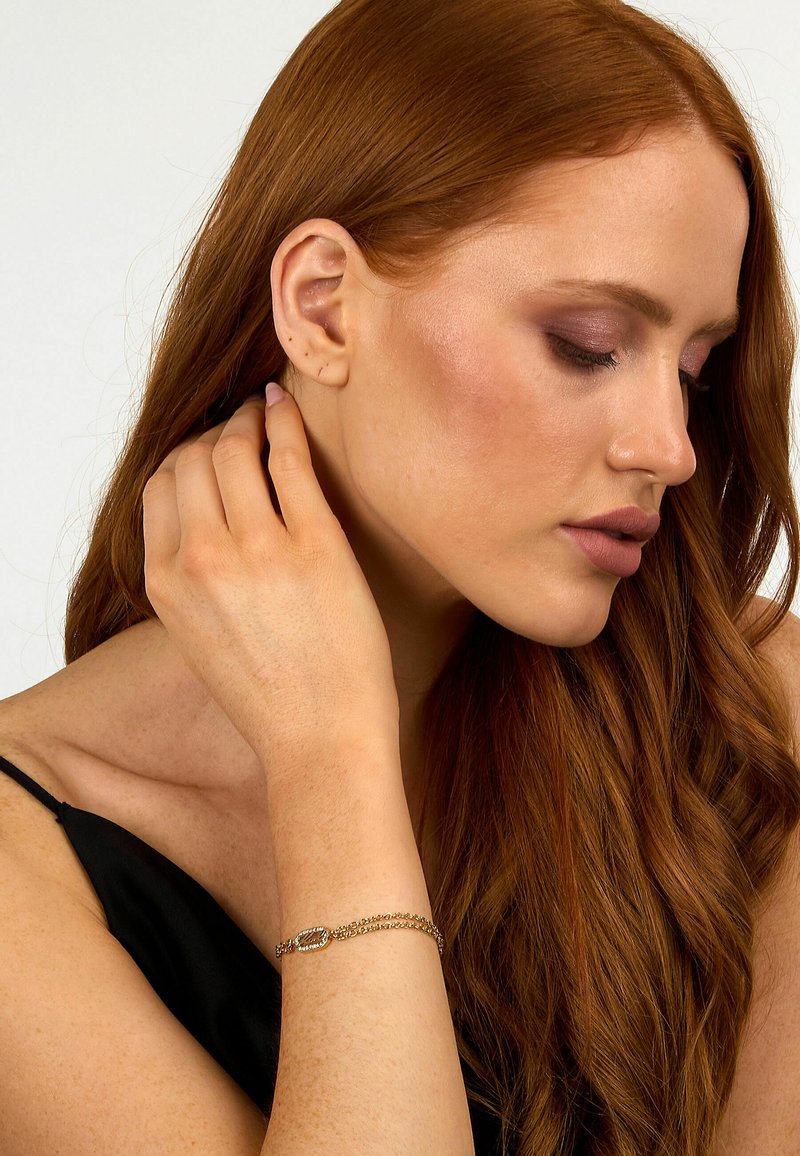 Guldtonad armband med länkdesign och diamantliknande detaljer, bärs på ett bar handled. Modellen har långt rött hår, slät hy och subtil sminkning.