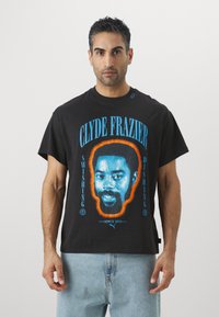 Sort bomulds T-shirt med et grafisk print af Clyde Frazier's ansigt i blå og orange. Rund hals, korte ærmer, afslappet pasform.