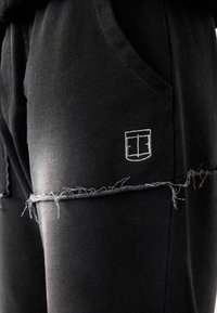 Schwarze Jogginghosen aus weichem Stoff, mit einem fransigen Saum, seitlichen Taschen und einem kleinen weißen gestickten Logo am Oberschenkel.