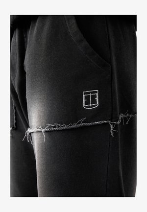 Black joggers made of soft fabric, featuring a distressed hem, side pockets, and a small white embroidered logo on the thigh.