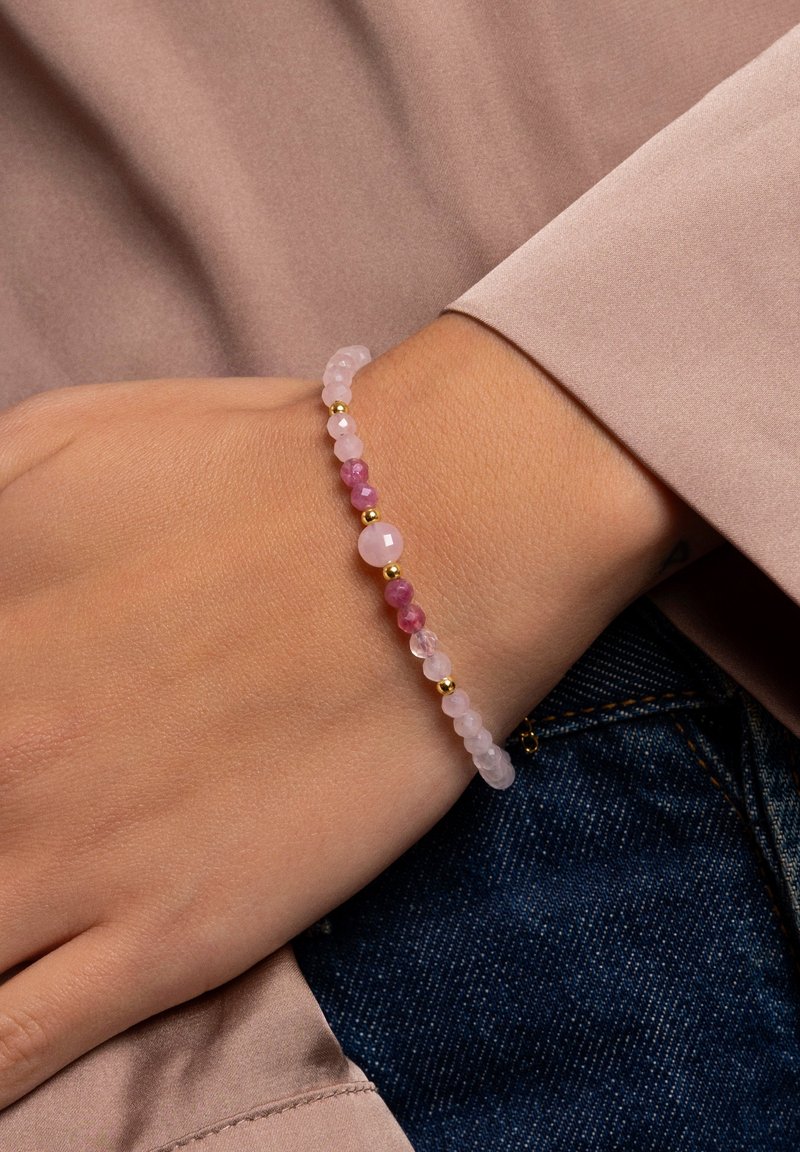 Main portant un bracelet de perles avec des pierres rondes roses et dorées, reposant sur un tissu beige et un jean en denim bleu.