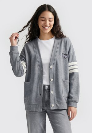 Ung kvinde smiler, iført en grå knap-op cardigan med hvide striber på det ene ærme og forlommer over en hvid skjorte og grå bukser.