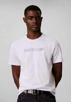 Άνδρας που φοράει λευκό t-shirt Calvin Klein και μαύρη ζώνη με σκούρο τζιν, στέκεται μπροστά σε απλό ανοιχτό γκρι φόντο.