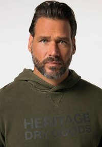 Olijfgroene hoodie met een gestructureerde afwerking, voorzien van een trekkoordkap en "HERITAGE DRY GOODS" in donkergrijs op de voorkant gedrukt.