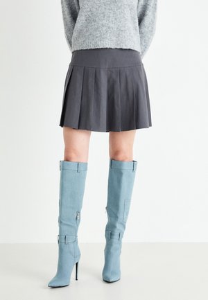 Femme portant une jupe plissée grise, un pull gris duveteux et des bottes hautes à talons bleu clair, debout sur un sol blanc.
