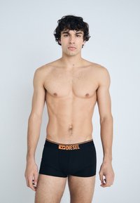 Boxer-briefs noirs avec un waistband orange affichant le logo "DIESEL". Le tissu est lisse et épouse étroitement le corps. Un modèle masculin pose sur un fond clair.