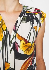 Femme portant une robe sans manches à motifs floraux avec des pétales jaunes, orange, rouges, verts, blancs et noirs et un décolleté en V.