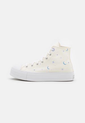 Białe sneakersy za kostkę z niebieskimi wzorami półksiężyców i gwiazd, białe sznurówki i gruba biała gumowa podeszwa, widok z boku.