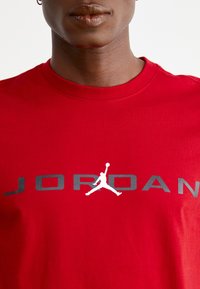 Rød bomulds T-shirt med rund hals, der har en stor grå "JORDAN" tekst og et hvidt silhuetlogo af en springende figur.