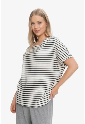 Kobieta ubrana w biało-granatowy pasiasty krótki top z krótkim rękawem i szare spodnie, pozująca z jedną ręką na biodrze na białym tle.