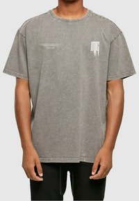 Grå oversized bomulds T-shirt med korte ærmer, rund hals og diskrete grafiske detaljer på brystet. Teksturfinish og slidseret udseende.