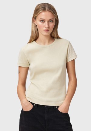 Ung kvinde med langt blondt hår iført en simpel cremefarvet t-shirt og sorte bukser, hænderne i lommerne, neutral ansigtsudtryk, lys grå baggrund.