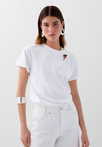 Hvid T-shirt lavet af let bomuld, med en knudefront ved skulderen, en skrå søm og en lille udsparing.