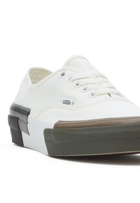 White canvas low-top sneakers with rubber accents. Features round laces, metal eyelets, and a textured green and brown sole.