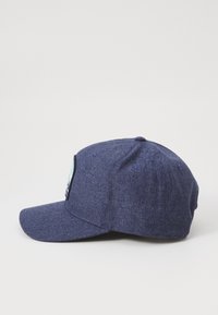 Marineblå caps med en litt buet skjerm, laget av teksturert stoff, med en frontlapp med grafisk detaljer. Bakre ventilasjons hull synlige.