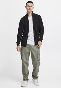 Schwarze gerippte Zip-Up-Strickjacke über einem grauen T-Shirt, kombiniert mit olivfarbenen Cargo-Hosen mit großen Taschen. Getragen mit weißen Sneakers.