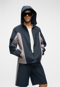 Άνδρας με μπλε ναυτικό και γκρι μπουφάν με κουκούλα και φερμουάρ, λευκό μπλουζάκι, κοντά παντελόνια που ταιριάζουν, γυαλιά ηλίου και ασημένιο αλυσίδα για λαιμό, στέκεται με τα χέρια στις τσέπες.